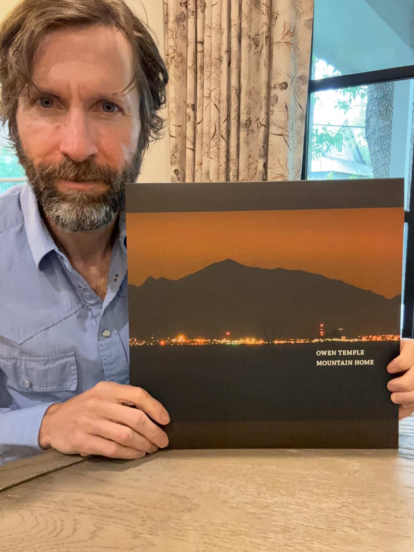 Man with beard in a blue shirt sits at a table, holding a landscape photo book showing a sunset over mountains and city lights (text: Owen Temple Mountain Home).