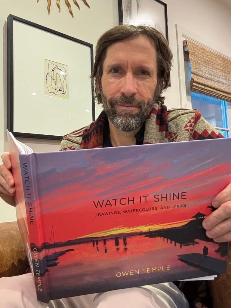Owen Temple holding up a copy of Watch It Shine, its cover a red and purple sunset over water
