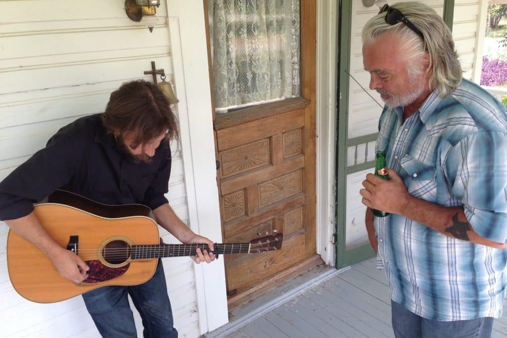 Owen Temple playing guitar on a porch while Hal Ketchum listens, D'Hanis, Texas, 2013